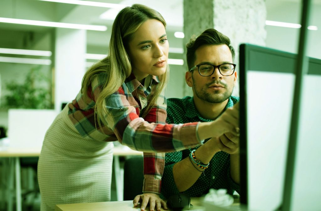 mulher loira de saia e blusa estampada, de pé, conversando com um rapaz sentado de topete e óculos, discutindo os tipos de arquiteturas de software